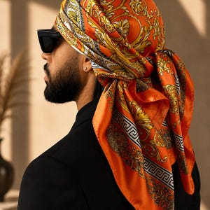 May include: A vibrant orange silk headscarf featuring an intricate gold and white baroque pattern with a black and white Greek key border. The square scarf is tied around the head, showcasing a glossy finish and detailed ornamental scrollwork designs.