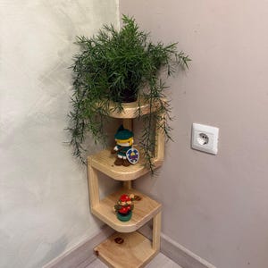 May include: A light-coloured wooden corner shelf with a zig-zag design featuring three tiers. The top tier holds a green leafy plant in a black pot, the middle tier holds a small crocheted figure with a shield, and the bottom tier holds a small figurine.