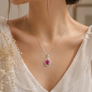 May include: A silver necklace featuring a delicate chain and a pendant with a central oval-shaped vibrant pink gemstone. The stone is surrounded by a decorative silver frame with four rounded petal-like loops, creating a floral-inspired design.