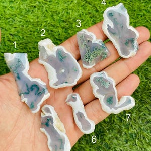 Puede incluir: Siete rodajas de ágata planas e irregulares numeradas del uno al siete. Cada piedra translúcida presenta un centro gris lavanda pálido con bordes blancos e inclusiones verdes distintivas similares al musgo en las superficies.