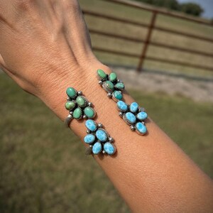 May include: A silver cuff bracelet featuring two clusters of oval-shaped stones. One cluster contains green turquoise-colored stones, while the other cluster features bright blue stones set in a decorative metal frame.