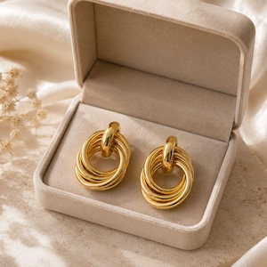 May include: A pair of gold-toned earrings, each featuring a design of multiple interlocking circular rings. The earrings have a polished, reflective finish and are presented inside a beige, rectangular, velvet-lined jewelry box.