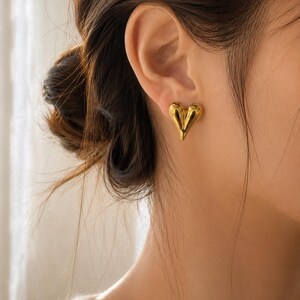 May include: A polished gold-tone stud earring shaped like a three-dimensional heart. The earring features a smooth, reflective metallic surface with a rounded, puffed design that sits flush against the earlobe, showcasing a bright yellow gold finish.