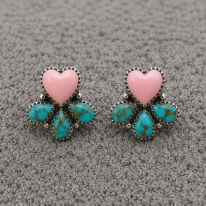 May include: A pair of stud earrings featuring a central smooth pink heart-shaped stone surrounded by four teardrop-shaped turquoise stones with brown matrix veining, all set in a silver-toned metal frame with a scalloped edge design.