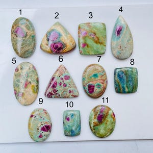 May include: Eleven polished cabochon gemstones arranged in rows, numbered one through eleven. These stones feature a mix of green, tan, and blue matrix with embedded magenta-colored inclusions in various oval, triangular, and rectangular shapes.