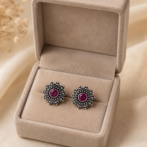 Puede incluir: Un par de pendientes de botón con una piedra central circular de color magenta rodeada por un marco plateado en forma de sol con pequeños detalles de cuentas, presentados en un joyero de terciopelo beige con tapa y forro suave.