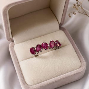 May include: A silver-toned ring featuring five oval-cut, deep pinkish-red gemstones arranged in a row. The stones are set in a prong setting, creating a continuous line of color across the band. The ring is displayed inside a velvet-lined box.