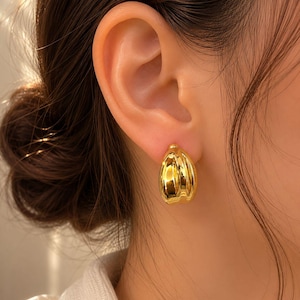 May include: A polished gold-tone hoop earring with a rounded, segmented design. The earring features a smooth, reflective metallic surface with vertical grooves that create a subtle, curved shape, securely fastened to the earlobe.