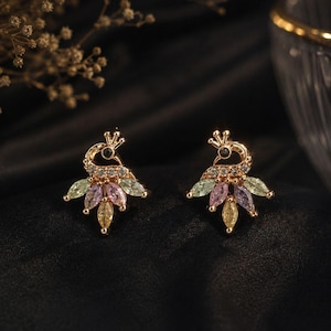 May include: A pair of gold-colored peacock earrings. Each earring features a peacock design with a crown, a black eye, and a fan of colorful gemstones in shades of green, pink, purple, and yellow. The earrings are set against a black background.