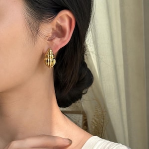 Peut inclure: Une paire de boucles d'oreilles en forme de coquillage dorées. Les boucles d'oreilles présentent des détails de crêtes et un motif en spirale. Elles sont portées sur l'oreille d'une personne aux cheveux foncés.