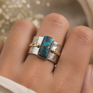 May include: A wide band ring featuring a rectangular turquoise stone with copper matrix veins. The band is crafted from hammered silver and accented with two thin, smooth gold-toned metal bands that frame the central stone setting.
