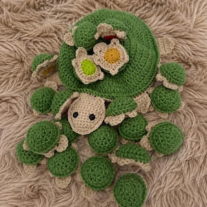 May include: A large crocheted green turtle with a tan head and black eyes, featuring a shell adorned with several smaller crocheted turtles in green and tan, along with small crocheted flower accents in yellow, green, and white colors.