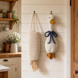 May include: A cream-colored crochet bag with a hanging loop and a beige plastic bag protruding from the bottom, next to a crochet goose figure featuring a white body, a yellow beak and feet, and a dark blue ribbon tied around its neck.