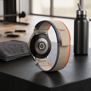 May include: A silver smartwatch with a beige, navy blue, and orange band. The watch is on a black surface next to a black water bottle and a gray towel. The watch band is made of a textured fabric.
