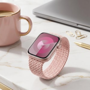 Puede incluir: Un reloj inteligente de oro rosa con una correa trenzada rosa. La esfera del reloj muestra un diseño degradado rosa con un reloj. Una taza rosa con borde dorado y un portátil están en el fondo.