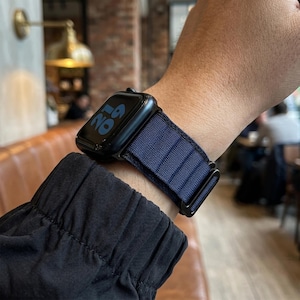 Op de afbeelding: Een zwarte smartwatch met een marineblauwe band wordt om een pols gedragen. De wijzerplaat toont de tijd in blauwe cijfers. De band heeft een getextureerd uiterlijk en een zwarte sluiting.