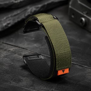 Könnte beinhalten: Ein olivgrünes und schwarzes Uhrenarmband aus gewebtem Nylongewebe. Das Armband verfügt über eine schwarze Metallschnalle und Anschlussbeschläge sowie eine kleine, leuchtend orangefarbene Stofflasche am Ende mit einem schwarzen Dreieckssymbol.