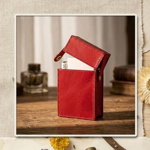 May include: A rectangular red leather case with a hinged lid that is partially open, revealing a white rectangular object inside. The case features visible stitching along the edges and a small gold-toned metal snap fastener on the side.