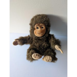 May include: A plush toy shaped like a young ape with dark brown shaggy fur covering its body and head. The face, hands, and feet are made of a smooth, light tan material. It has dark, round eyes and a small, gentle smile on its face.
