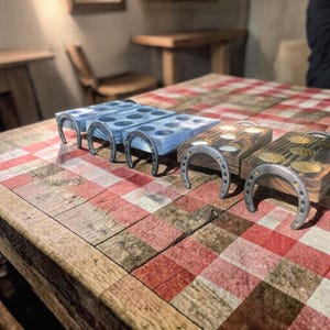 Puede incluir: Soportes de madera para bebidas con asas metálicas en forma de herradura. Los soportes están pintados en tonos azules y marrones, con recortes circulares para sujetar vasos. Los soportes se exhiben sobre una mesa a cuadros rojos y blancos.