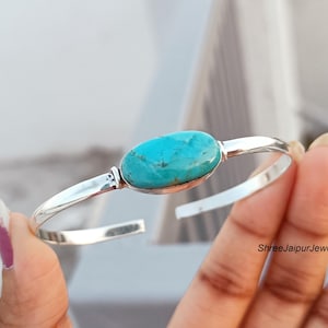Könnte beinhalten: Ein silbernes Armband mit einem zentralen, ovalen Türkis-Edelstein mit natürlicher Maserung. Das polierte Metallband hat ein offenes Design, und der Text ShreeJaipurJewels92 ist auf der unteren rechten Seite des Schmuckstücks zu sehen.