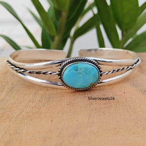 May include: A silver cuff bracelet with a turquoise oval stone at the center. The bracelet has a twisted rope design on the sides and is displayed on a wooden surface. The text "SilverJewels26" is visible.