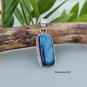 Op de afbeelding: Een rechthoekige hanger met een gepolijste blauwe labradoriet edelsteen in een zilveren zetting met een metalen oogje aan de bovenkant. De steen vertoont iriserende blauwe flitsen en natuurlijke barsten. De tekst SilverJewels26 is zichtbaar.