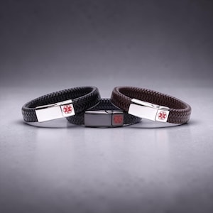 May include: Three braided leather medical alert bracelets in black, dark brown, and medium brown. Each bracelet has a silver or dark grey clasp with a red medical symbol. The bracelets are displayed on a grey surface.
