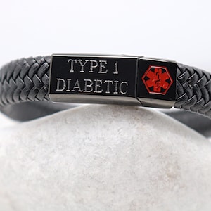 Puede incluir: Pulsera de cuero trenzado negro con una placa de metal negro que dice "TYPE 1 DIABETIC" y un símbolo de alerta médica rojo. La pulsera está sobre una piedra blanca.