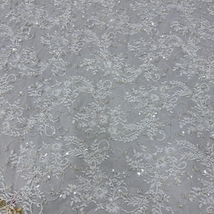 Op de afbeelding: Close-up van witte kanten stof met bloemen- en bladerpatronen. De stof is versierd met kleine, glanzende pailletten en kleine parelachtige kralen, wat een delicate en getextureerde oppervlakte creëert. Het kantontwerp is ingewikkeld en gedetailleerd, voor bruids- of formele kleding.