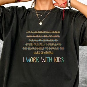 Peut inclure: Un t-shirt gris anthracite à manches courtes avec le texte "I'M A SCIENTIST PRACTITIONER WHO APPLIES THE NATURAL SCIENCE OF BEHAVIOR TO SYSTEMATICALLY MANIPULATE THE ENVIRONMENT TO IMPROVE THE LIVES OF OTHERS" et "I WORK WITH KIDS" en couleurs.