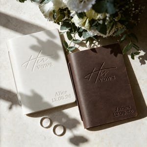 May include: A cream-colored notebook embossed with Her Vows and Alice 12.06.26, and a dark brown notebook embossed with His Vows and James 12.06.26, accompanied by two silver-toned metal rings positioned below the books on a light surface.