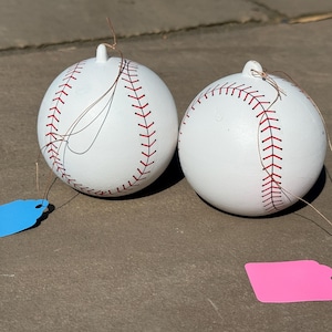 May include: Two white baseball ornaments with red stitching details. Each ornament has a small loop at the top and a tag attached with string. One tag is blue, and the other is pink.