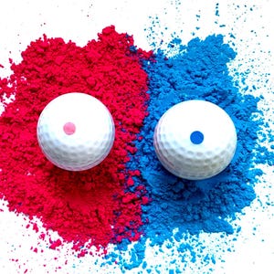 May include: Two white golf balls, each with a colored dot, are surrounded by vibrant red and blue powder. The golf balls are positioned on a white background, creating a striking contrast. The image suggests a gender reveal or a color-themed event.