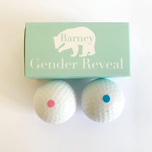 May include: A light mint green rectangular box with a white bear silhouette and the text Barney Gender Reveal, accompanied by two white golf balls, one featuring a small pink dot and the other featuring a small blue dot on the surface.