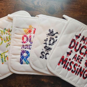May include: Four white quilted pot holders with various text and duck graphics. One says "My Ducks are Thriving" in colorful letters. Another says "Ducks are Rad" in rainbow colors. The third says "Ducks are So" in black. The last says "My Ducks are Missing" in red and black plaid.
