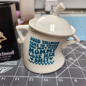 May include: A white ceramic sugar bowl with a lid and two handles. The bowl has blue text that reads "GOOD THINGS COME TO THOSE WHO DON'T MAKE MOMMY LOSE HER SHIT". The bowl is on a gray cutting mat.