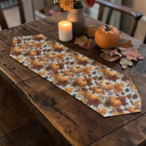 May include: A white table runner featuring a repeating pattern of orange pumpkins, brown acorns, and multicoloured autumn leaves. The runner is laid flat on a dark wooden table, accompanied by a lit white candle and a small orange pumpkin.