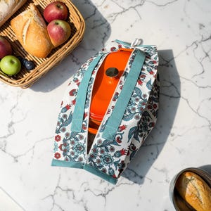 Könnte beinhalten: Ein orangefarbener emaillierter gusseiserner Bräter befindet sich in einer floral gemusterten Tragetasche mit türkisfarbenen Trägern. Die Tasche hat einen weißen Hintergrund mit einem blauen und roten Blumenmuster. Ein Korb mit Brot und Äpfeln befindet sich im Hintergrund.