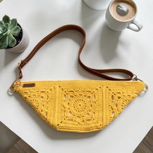 May include: A yellow crochet fanny pack with a brown leather strap and gold-colored hardware. The bag features a detailed square pattern in the center and is displayed on a white surface. The bag is a stylish accessory.