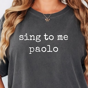 May include: A charcoal gray short-sleeve t-shirt featuring the white text "sing to me paolo" printed in a typewriter-style font across the chest. The garment has a crew neckline and a relaxed fit, paired with blue denim jeans.