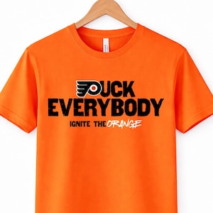 May include: Bright orange t-shirt with the text "DUCK EVERYBODY" in bold black letters. The shirt also features a logo and the words "IGNITE THE ORANGE" in white. The shirt is hanging on a wooden hanger.