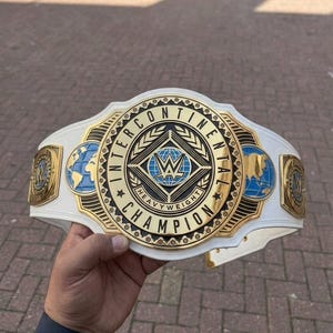 Pode incluir: Um cinturão de campeonato Intercontinental branco e dourado com detalhes azuis. O cinturão apresenta o logotipo da WWE e as palavras "Intercontinental Champion" em letras douradas. O cinturão tem uma correia branca.