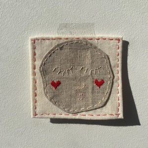 May include: A square piece of off-white fabric featuring a circular beige linen patch embroidered with a sleeping face. The face has closed eyes, a small mouth, and two red heart-shaped cheeks. The patch is outlined with white stitching.