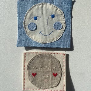 May include: Two square fabric patches featuring embroidered faces. The top patch is light blue denim with a cream circular face, blue eyes, blue cheeks, and a smile. The bottom patch is cream fabric with a tan circular face, closed eyes, and red hearts.