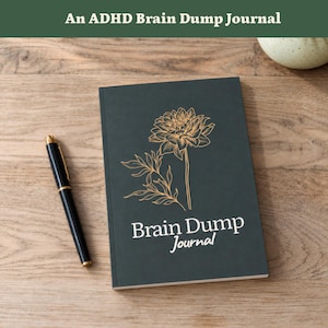 Può includere: Un diario grigio antracite scuro con un'illustrazione floreale a linee dorate sulla copertina. Il testo "Brain Dump Journal" è stampato in bianco sotto il fiore. Una penna a sfera nera con dettagli dorati è posizionata a sinistra del diario.