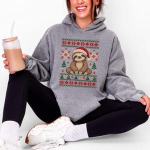 May include: A heather gray hooded sweatshirt featuring a central graphic of a brown sloth wearing a red Santa hat, surrounded by a festive pattern of green Christmas trees, red snowflakes, and geometric shapes in red and green.