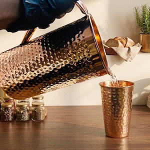 Puede incluir: Una jarra de cobre vierte agua en una taza de cobre a juego. Ambos artículos tienen una textura martillada. La jarra es grande, con un asa. La taza es cilíndrica. La escena está ambientada sobre una superficie de madera, con otros artículos de cocina al fondo.