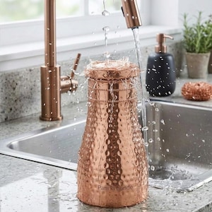 May include: A copper-colored hammered metal pitcher with water splashing over the top. The pitcher is near a copper faucet and a stainless steel sink. A black soap dispenser and a copper scrub are in the background. The text "Pani ki bunde" is visible on the pitcher.