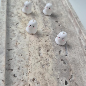 May include: Four small, white, ghost-shaped beads with smooth surfaces. Each bead features a simple printed face with two black circular eyes and two small pink circular cheeks. Each ghost bead has a small hole at the top for threading purposes.